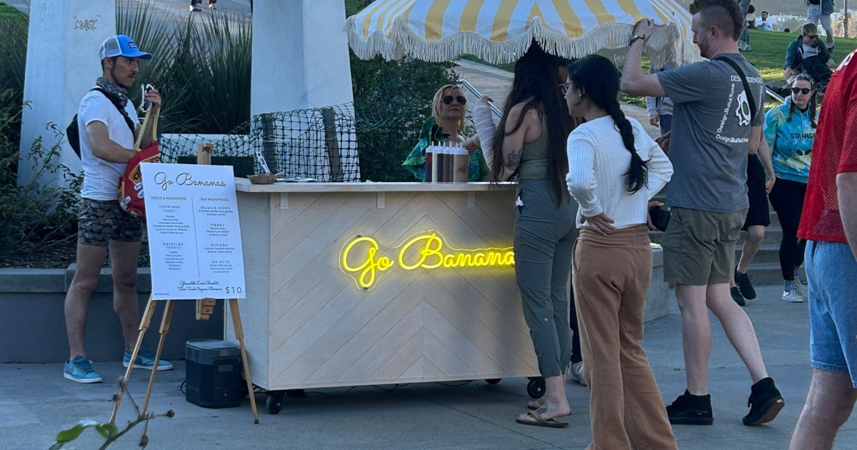 A chocolate-covered banana on a stick is drizzled with peanut butter and sprinkled with crushed nuts, held in a small brown paper tray.