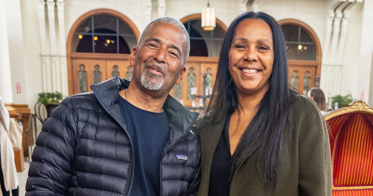 A smiling man in a black puffer jacket and jeans stands next to a smiling woman in a green coat in an ornately decorated room with arched doorways in the background.