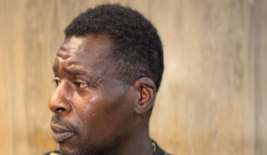 A man with short curly hair and a mustache looks to the left, wearing a black shirt with gold and white geometric patterns, standing against a blurred background.