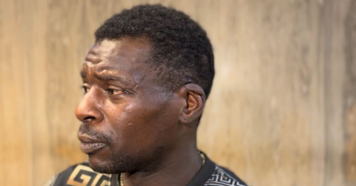 A man with short curly hair and a mustache looks to the left, wearing a black shirt with gold and white geometric patterns, standing against a blurred background.