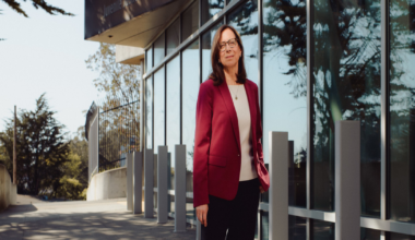 A woman with shoulder-length brown hair and glasses stands confidently in a red blazer outside the Juvenile Justice Center building.