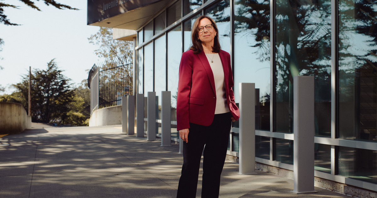 A woman with shoulder-length brown hair and glasses stands confidently in a red blazer outside the Juvenile Justice Center building.