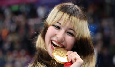 A young woman with blonde and brown hair smiles widely while biting a gold Olympic medal, holding a flag and wearing a sequined outfit.