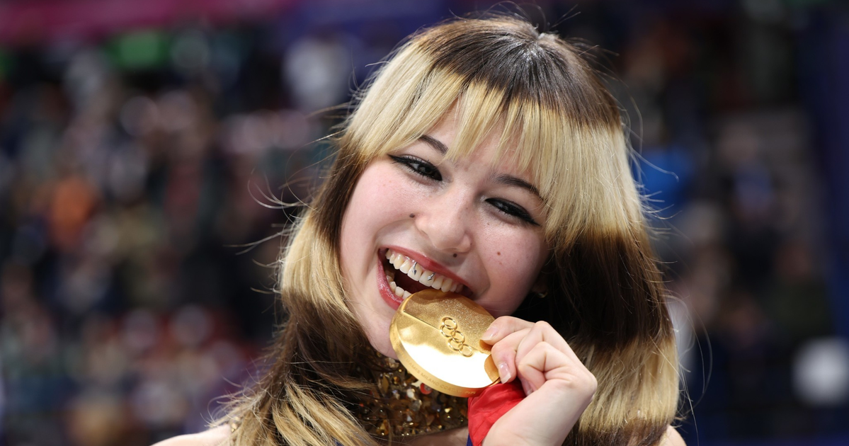A young woman with blonde and brown hair smiles widely while biting a gold Olympic medal, holding a flag and wearing a sequined outfit.