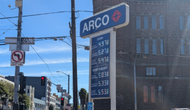 An ARCO gas station sign lists prices for cash and debit/credit, with a no left turn sign and a 20 mph speed limit near an intersection with pedestrians.