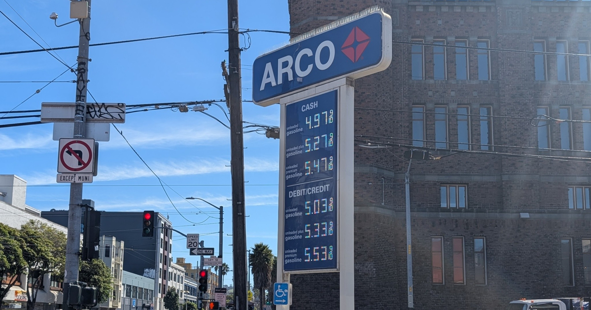 An ARCO gas station sign lists prices for cash and debit/credit, with a no left turn sign and a 20 mph speed limit near an intersection with pedestrians.