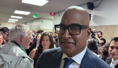 A man in a suit and large glasses stands in a crowded hallway with people talking and cameras in the background.