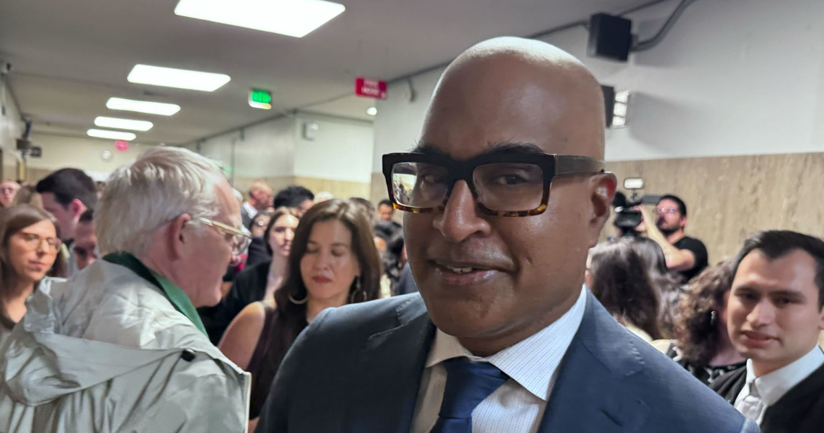 A man in a suit and large glasses stands in a crowded hallway with people talking and cameras in the background.