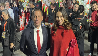 A man in a suit and red tie stands beside a smiling woman in a red cape with a ribbon during a busy, colorful street festival with many people.
