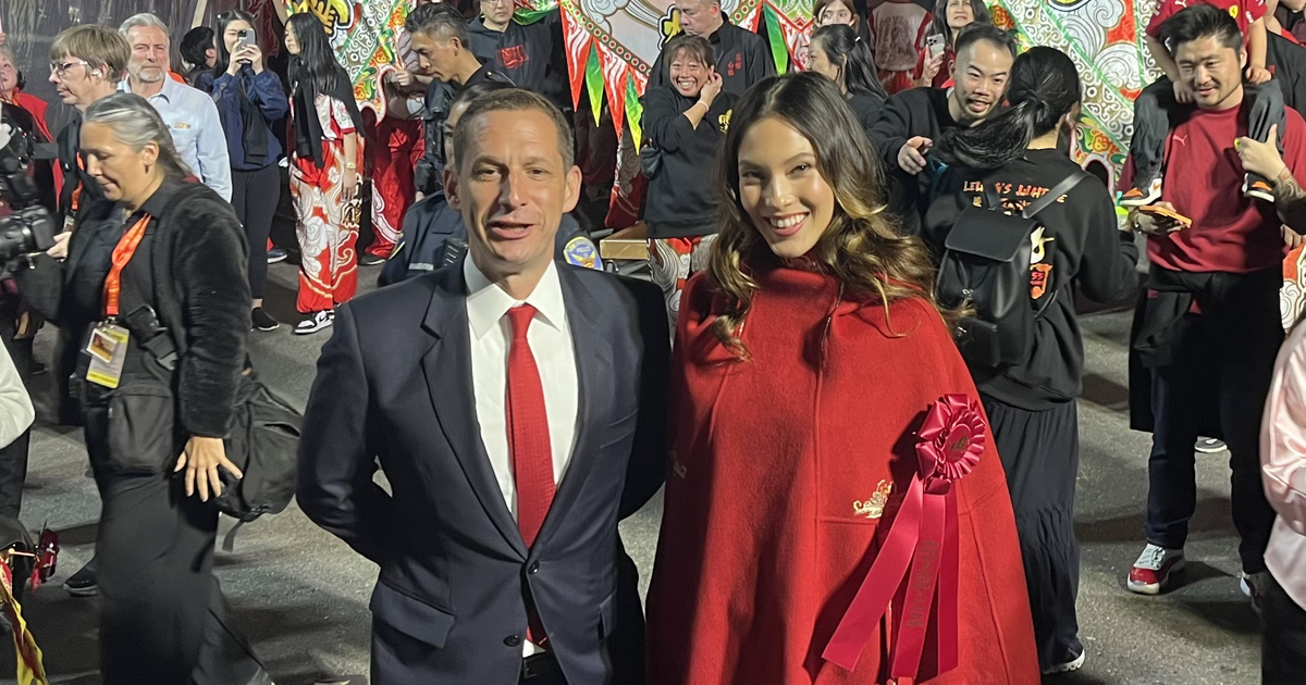 A man in a suit and red tie stands beside a smiling woman in a red cape with a ribbon during a busy, colorful street festival with many people.