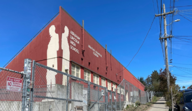 A red building labeled Abraham Lincoln High School and Sebastian Scandura Jr. Stadium stands behind a chain-link fence along a sunny sidewalk with parked cars and utility poles.