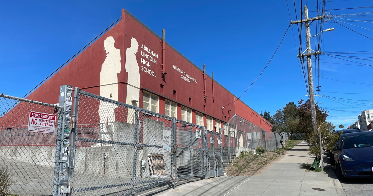 A red building labeled Abraham Lincoln High School and Sebastian Scandura Jr. Stadium stands behind a chain-link fence along a sunny sidewalk with parked cars and utility poles.