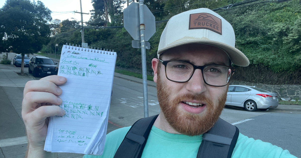 A man with glasses and a beard wears a light green shirt and tan hat while holding a notepad with tally marks at Cesar Chavez and Douglass streets.