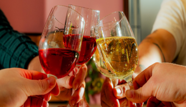 Three people smiling indoors, two seated holding glasses of wine, one standing behind them with a glass of red wine, against an orange and pink wall.