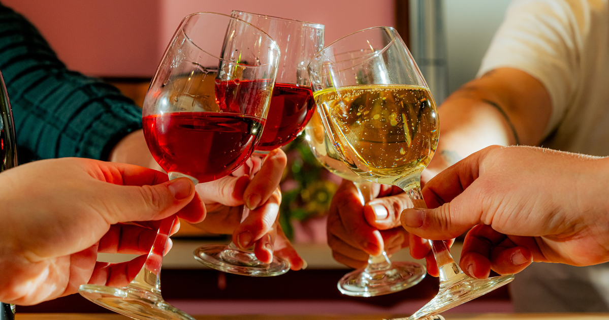 Three people smiling indoors, two seated holding glasses of wine, one standing behind them with a glass of red wine, against an orange and pink wall.