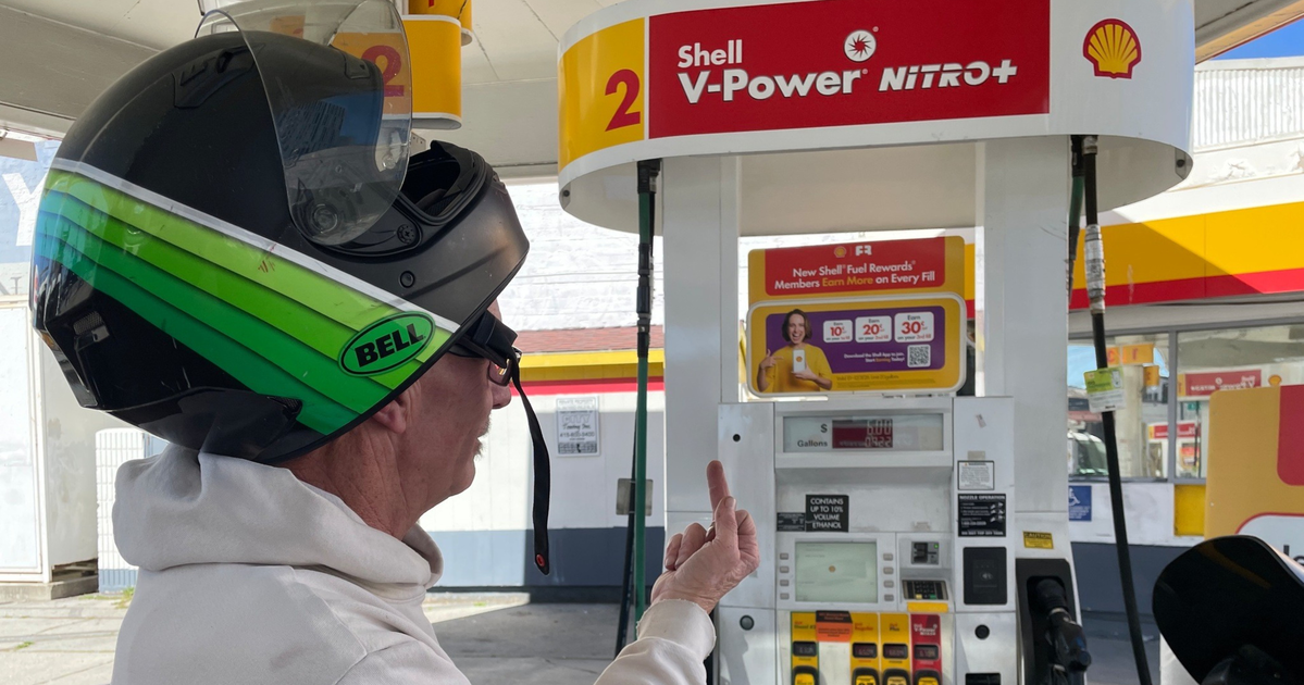 A person wearing a white hoodie and a black helmet with green stripes stands at a Shell V-Power Nitro+ gas pump, raising their middle finger.