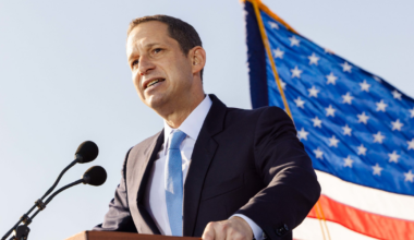 A man in a suit and light blue tie speaks at a podium with two microphones, with a large American flag waving in the background.