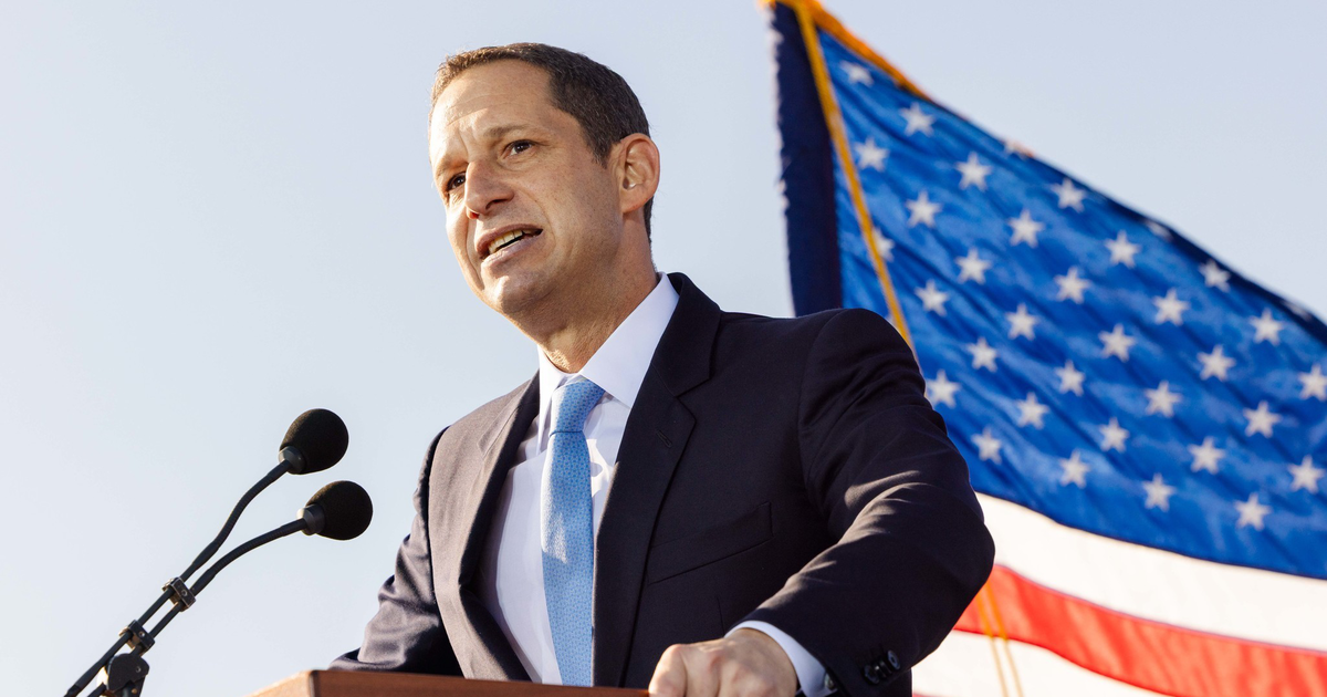 A man in a suit and light blue tie speaks at a podium with two microphones, with a large American flag waving in the background.