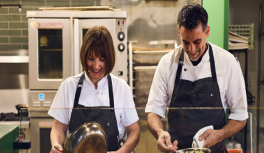 A man and woman in white shirts and dark aprons smile while holding a small child dressed in green pajamas, standing in a warmly lit room.