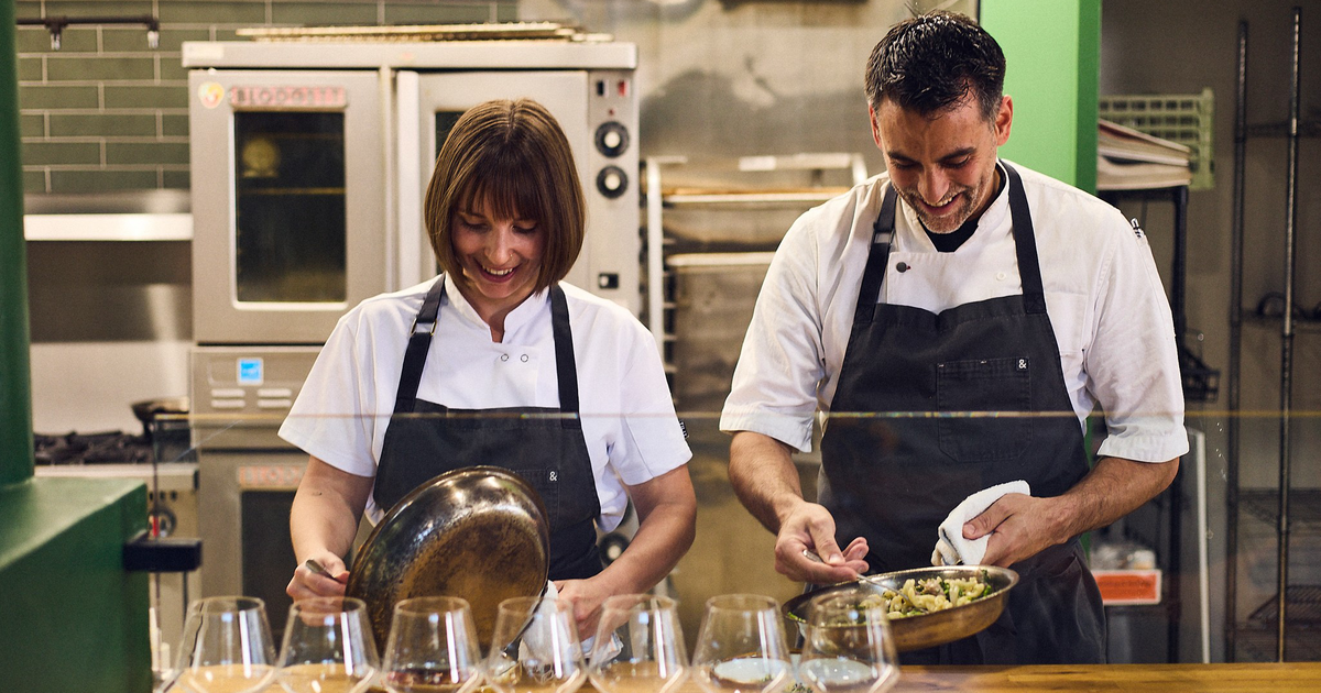 A man and woman in white shirts and dark aprons smile while holding a small child dressed in green pajamas, standing in a warmly lit room.