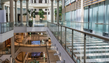 A spacious, multi-level indoor area with glass railings, large columns, plants, and natural light streaming through arched windows and a glass ceiling.
