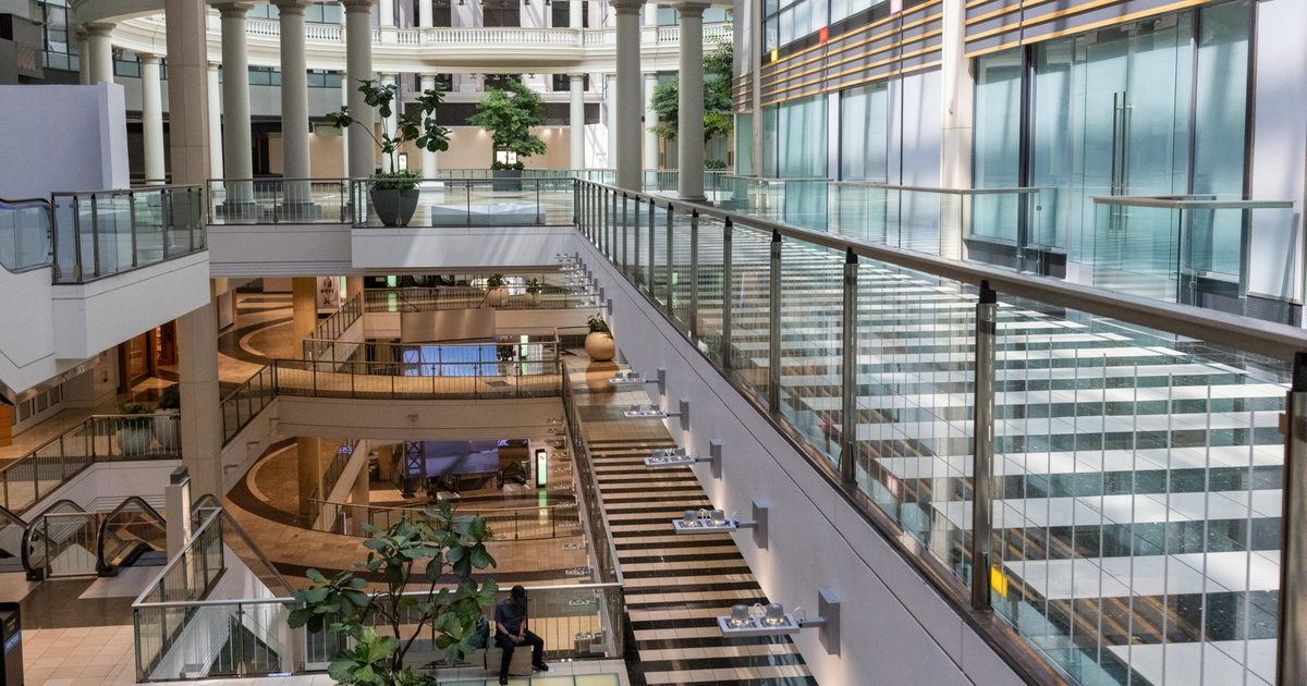 A spacious, multi-level indoor area with glass railings, large columns, plants, and natural light streaming through arched windows and a glass ceiling.