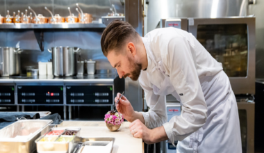 A chef in a white apron leans on a counter in a modern kitchen while two other chefs work preparing food in the background.