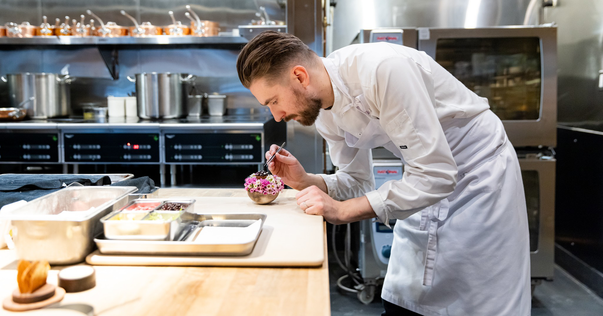A chef in a white apron leans on a counter in a modern kitchen while two other chefs work preparing food in the background.