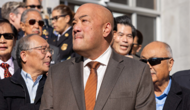 A group of men, mostly wearing suits and sunglasses, gather outside in daylight, with one bald man in a brown suit and orange tie standing prominently in front.