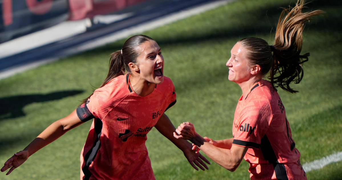 Two female soccer players in orange jerseys celebrate enthusiastically on the field, holding hands and smiling broadly.