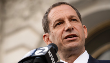 A man in a suit and tie speaks into multiple microphones labeled KTVU, NBC Bay Area, and another, with a blurred building in the background.