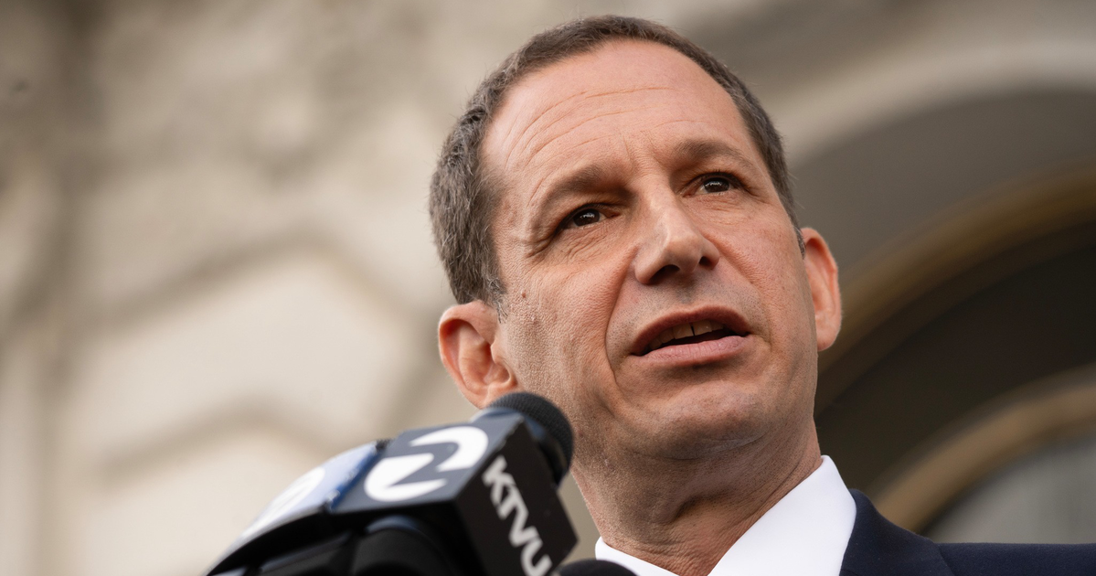 A man in a suit and tie speaks into multiple microphones labeled KTVU, NBC Bay Area, and another, with a blurred building in the background.