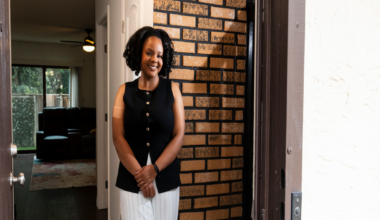 A smiling woman with curly hair wears a sleeveless black vest and light-colored pants, standing inside near a brick wall and white door.