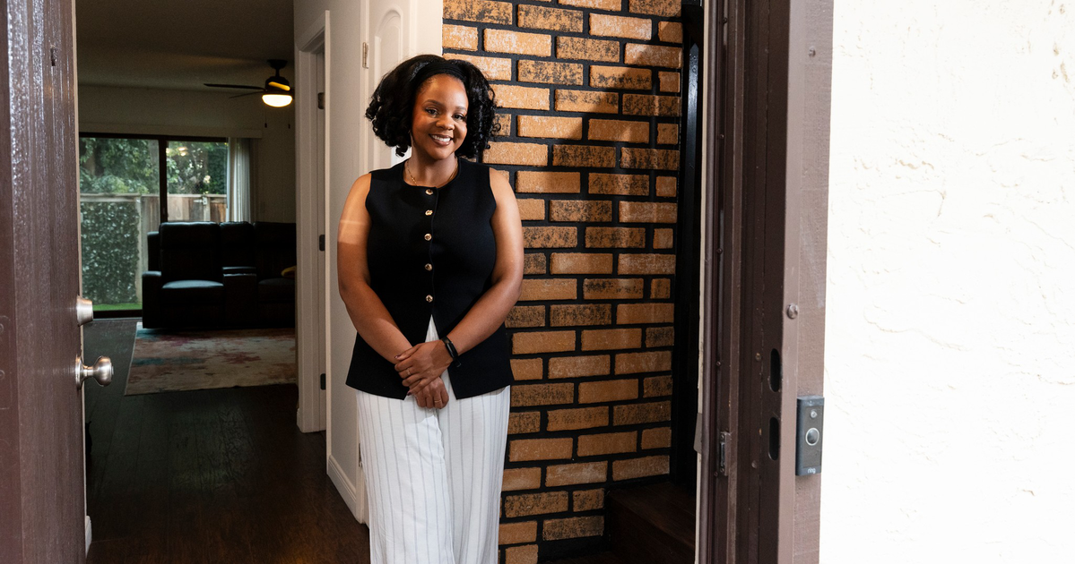 A smiling woman with curly hair wears a sleeveless black vest and light-colored pants, standing inside near a brick wall and white door.