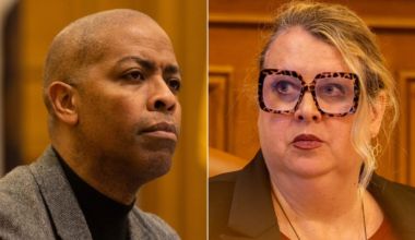 A man in a gray jacket and black turtleneck looks serious, while a woman with large tortoiseshell glasses and a dark blazer looks to the side.
