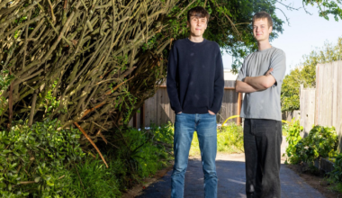 Two young men stand on a paved path between dense green bushes and a wooden fence, with one wearing a black sweater and jeans and the other in a gray shirt and black pants.