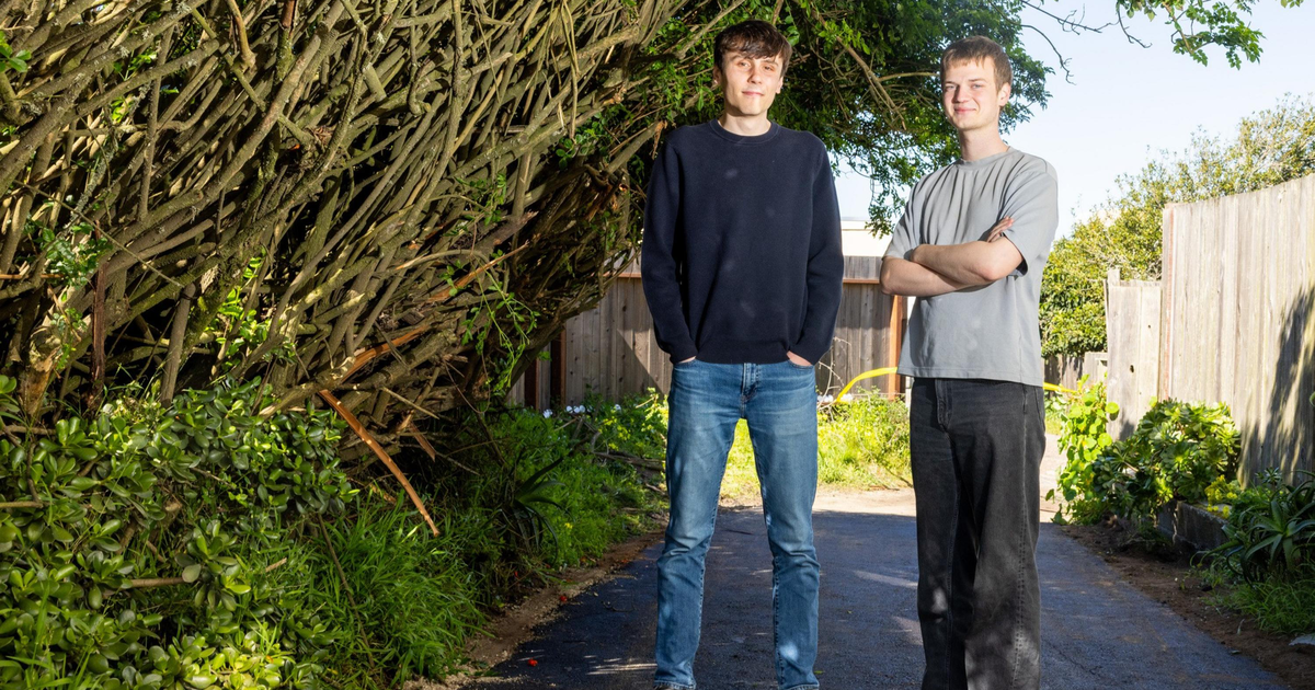 Two young men stand on a paved path between dense green bushes and a wooden fence, with one wearing a black sweater and jeans and the other in a gray shirt and black pants.