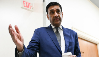 A man in a blue suit and patterned tie holds a coffee cup in one hand and gestures with the other while standing indoors near a wooden door and an exit sign.