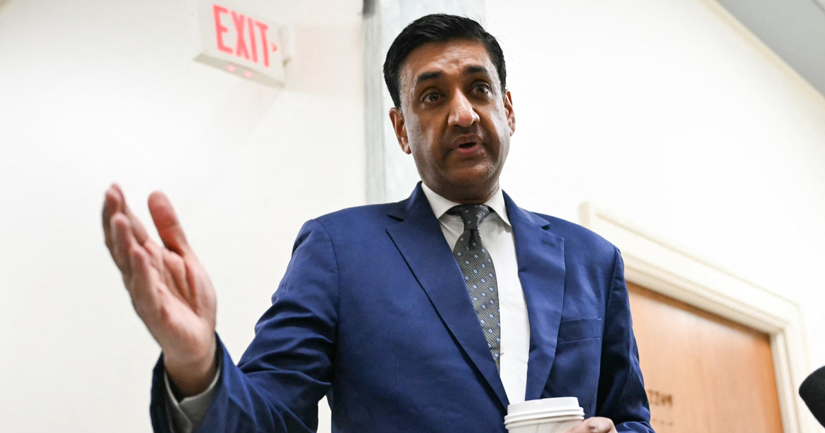 A man in a blue suit and patterned tie holds a coffee cup in one hand and gestures with the other while standing indoors near a wooden door and an exit sign.