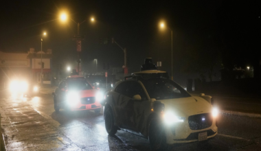 A white car with a rooftop sensor drives ahead of a police car with bright headlights on a wet street at night under streetlights.