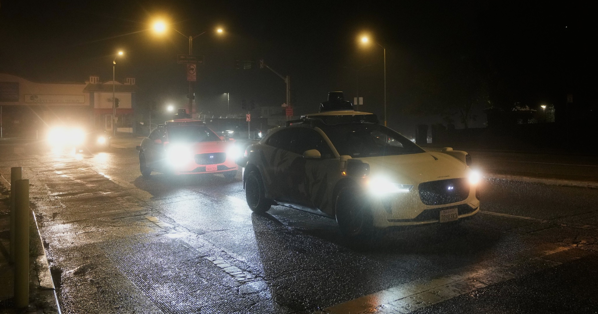 A white car with a rooftop sensor drives ahead of a police car with bright headlights on a wet street at night under streetlights.