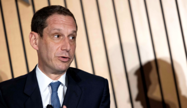A man in a dark suit and blue tie gestures with his right hand while speaking, casting a clear shadow on a wooden slatted wall behind him.