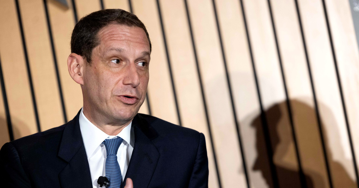A man in a dark suit and blue tie gestures with his right hand while speaking, casting a clear shadow on a wooden slatted wall behind him.