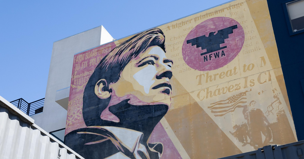 A large mural of a young man looking upward with the NFW logo and faded text about Chavez and farm workers is painted on a building.