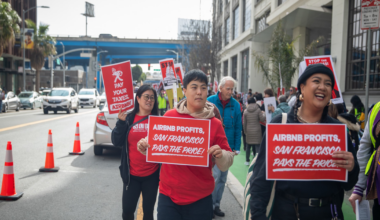 People are marching on a city street holding signs that read, "AIRBNB PROFITS, SAN FRANCISCO PAYS THE PRICE!" and "PAY YOUR TAXES, AIRBNB."