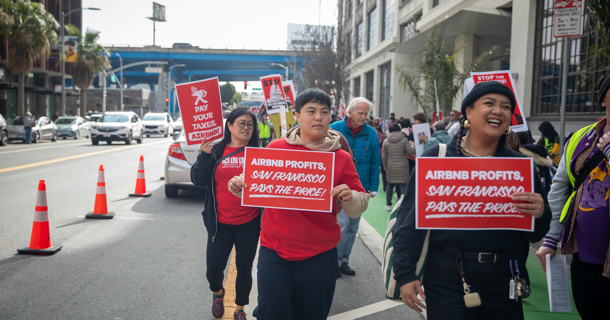 People are marching on a city street holding signs that read, "AIRBNB PROFITS, SAN FRANCISCO PAYS THE PRICE!" and "PAY YOUR TAXES, AIRBNB."