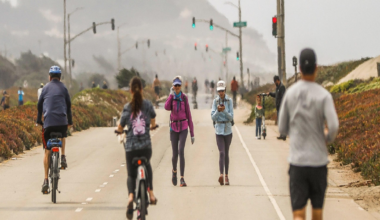 A road with cyclists, walkers, and a runner, with traffic lights overhead and foggy hills in the distance.