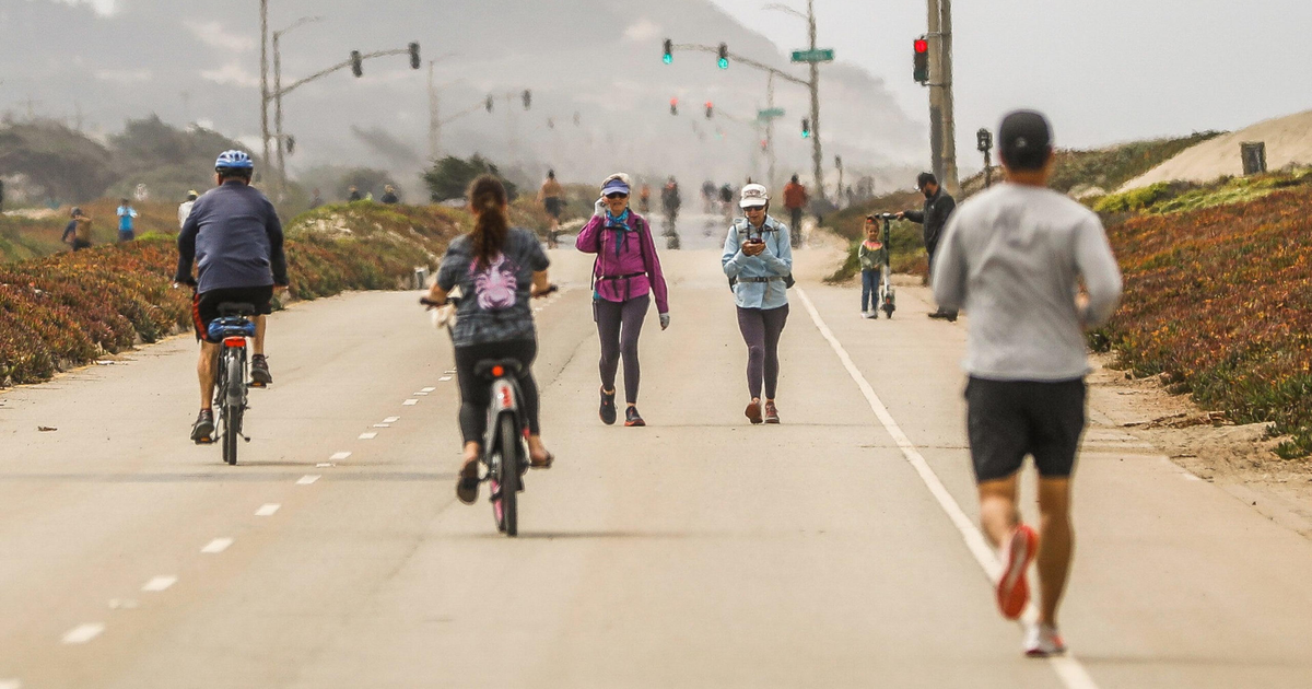 A road with cyclists, walkers, and a runner, with traffic lights overhead and foggy hills in the distance.
