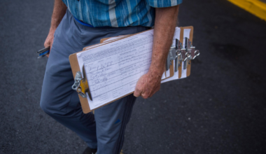 An older person in a blue plaid shirt and gray pants holds several clipboards with papers, standing on asphalt near a yellow curb.