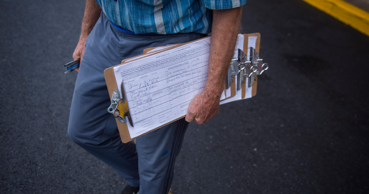 An older person in a blue plaid shirt and gray pants holds several clipboards with papers, standing on asphalt near a yellow curb.